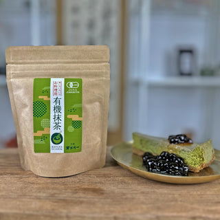 Zwei Objekte stehen auf einem antiken Holztisch. Links im Bild ist eine Packung Bio-Matcha zum Backen. Rechts im Bild ist ein goldener Teller mit einem Stück Matcha-Chesecake, garniert mit einem Kompott aus schwarzen Beeren