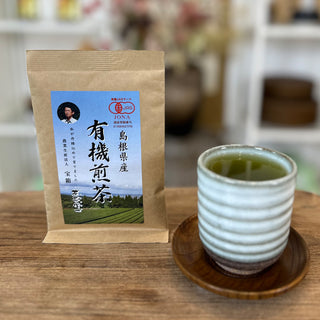 Bio-Sencha Takabarako loser Tee in einer Papierverpackung, daneben eine Teetasse namens Haru mit grünem Tee, auf einem Holztisch vor unscharfem Hintergrund.