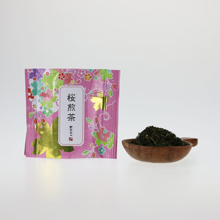 Eine rosarote Packung Sakura Sencha steht vor weißem Hintergrund. Rechts daneben ist ein Holzgefäß, gefüllt mit dem Sakura Sencha Tee, dem rote und rosarote Kirschblüten beigemengt sind.