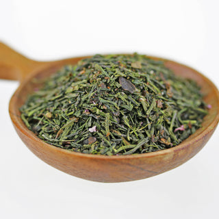 Eine Nahaufnahme von einer Portion Sakura Sencha in einem Holzgefäß. Man sieht die dunkelgrünen Teeblätter, die länglich geformt sind, ähnlich wie Tannennadeln. Dazwischen gemischt sind rosarote und violette kandierte Kirschblüten.