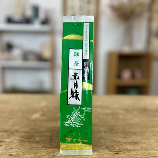 Eine längliche Packung "Satsuki Midori Sencha" in grün, mit einer weißen Zeichnung eines japanischen Schreins und japanischen Schriftzeichen auf der Vorderseite, steht auf einer hellen Holzplatte. 