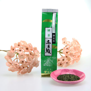 Vor einem weißen Hintergrund sieht man eine Packung "Satsuki Midori Sencha". Die Packung ist lang und grün, mit goldenen Elementen, japanischen Schriftzeichen und einer weißen Zeichnung verziert, die das Dach eines japanischen Schreins zeigt. Im Hintergrund sieht man rosarote Blüten. Vorne rechts im Bild ist ein Tellerchen in Pink, gefüllt mit Teeblättern.