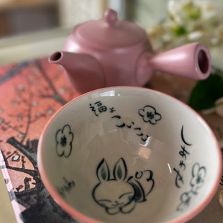 Eine "kawaii Bunny"-Keramikschale mit einem niedlichen weißen Hasen und weißen Blütenmuster, neben einer rosaroten Kyusu-Teekanne namens "Ichigo" auf einem dekorativen Untergrund mit Kirschblütenmotiv. Im Hintergrund sind unscharf einige Blumen zu sehen.