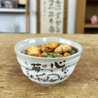 Die Schale "Neko" steht auf einem Holztisch, gefüllt mit bunten japanischen Snacks. Man erkennt das niedliche Katzenmotiv und die japanische Kalligraphie auf der Vorderseite der Schale. im Hintergrund sind verschwommen eine Schriftrolle sowie ein Regal zu erkennen.