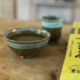 Eine mit grünem Tee gefüllte Schale "Sora" in den Farben hellblau und dunkelgrün steht auf einem Holztisch. Daneben liegt einen goldene Packung mit Genmai-Tee, auf der japanische Schriftzeichen zu sehen sind. Im Hintergrund des Bildes steht die Teetasse "Sora" in denselben Farben wie die Schale, Moosgrün und Himmelblau.