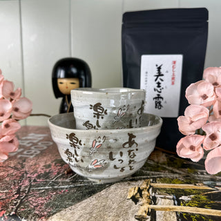 Die Schale "Usagi" und die dazugehörige Teetasse "Usagi" sind ineinander gestapelt. Sie sind beide hellgrau und mit niedlichen Häschen und japanischer Schrift dekoriert. Im Hintergrund steht eine japanische Holzpuppe, die ein Mädchen mit schwarzen Haaren darstellt, und rosarote Blumen sowie eine schwarze Packung "Izumo Taisha Sencha".