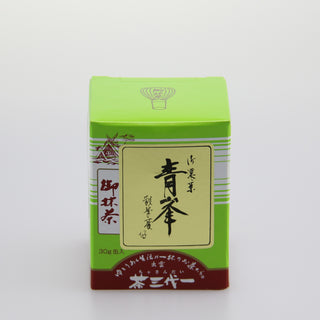 Eine grüne Packung Matcha mit der Bezeichnung "Seihou". Oben links ist eine Zeichnung eines japanischen Schreins zu sehen. Die Verpackung ist mit japanischen Schriftzeichen versehen und hat eine rotbraune Basis. Der Hintergrund ist neutral und weiß gehalten.