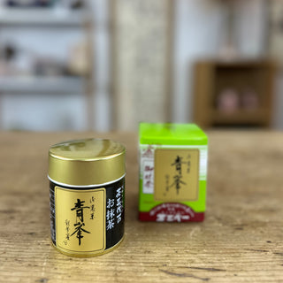 Im Vordergrund steht die Matcha-Dose "Seihou" mit goldenem Deckel und schwarzem Etikett. Dahinter befindet sich die grüne Verpackung des Matcha "Seihou" mit japanischen Schriftzeichen und einem braunroten unteren Bereich. Im Hintergrund sind unscharfe Regale und Dekorationsgegenstände zu erkennen, die eine gemütliche Atmosphäre andeuten.