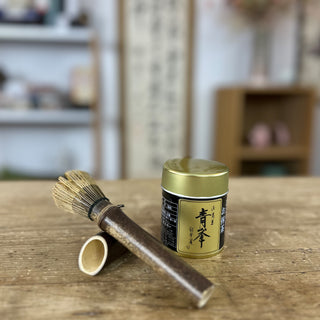 Im Vordergrund steht die Matcha-Dose "Seihou" mit goldenem Deckel und schwarzem Etikett. Daneben liegt der Matchabesen "Tenmoku schwarz" aus dunklem Holz. Im Hintergrund sind unscharfe Regale und Dekorationsgegenstände zu erkennen, die eine gemütliche Raumatmosphäre schaffen.