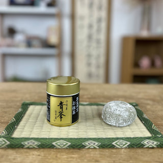 Die Matcha-Dose "Seihou" mit goldenem Deckel und schwarzem Etikett steht auf einer kleinen Tatami-Matte. Daneben liegt ein Mochi. Im Hintergrund sind unscharfe Regale und Dekorationsgegenstände zu sehen, die eine einladende Raumatmosphäre schaffen.