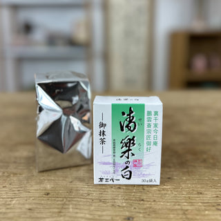 Der Matcha "Seiraku no Shiro" in einer weißen Verpackung mit hellgünem Strefen und schwarzen japanischen Schriftzeichen steht auf einer hellen Holzplatte. Dahinter ist der Inhalt zu sehen, eine silbrige Tüte.