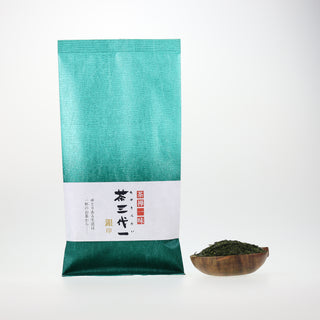 Eine Packung Sencha Gin in blau-grüner Verpackung mit Farbverlauf und japanischer Kalligraphie, daneben eine Holzschale mit grünen Teeblättern.