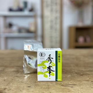 Eine Packung Bio-Matcha Shuhou steht auf einem Holztisch. Der innenliegende, silbrige Beutel befindet sich dahinter. Im Hintergrund sieht man eine japanische Kalligraphie