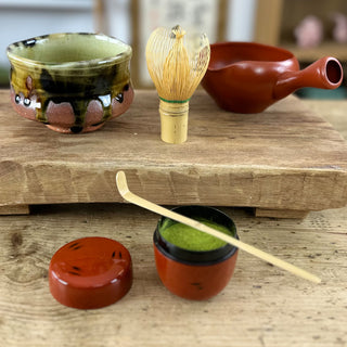 Auf einem Holztisch stehen mehrere Teezubehörteile, darunter eine grün-schwarze Matchaschale namens 'Bidoro', ein traditioneller Bambusbesen (Chasen), ein rotes Abkühlgefäß namens 'Yuzamashi' und eine Matcha-Teedose namens 'Negoro' mit traditionellem Muster in rot und schwarz, gefüllt mit grünem Matcha-Pulver. Der Deckel der Dose liegt daneben, und ein Bambus-Teelöffel ruht auf der Dose. Im Hintergrund sind ein Regal und eine japanische Schriftrolle zu erkennen.