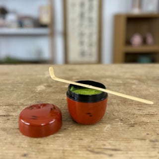 Matcha-Teedose namens 'Negoro' mit traditionellem Muster in rot und schwarz, gefüllt mit grünem Matcha-Pulver. Der Deckel liegt neben der geöffneten Dose, und ein Bambus-Teelöffel ruht auf der Dose. Im Hintergrund sind ein Regal und eine japanische Schriftrolle zu erkennen.