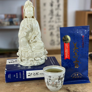 Vier Gegenstände befinden sich auf einem Holztisch. Im Vordergrund steht eine "Jizo"-Teetasse in hellgrau, gefüllt mit grünem Tee. Rechts daneben ist eine blaue Packung "Monde Mishiro Sencha" mit goldenen Schriftzeichen darauf zu sehen. Links davon liegt ein blaues Buch, auf dem eine buddhistische Statue aus weißer Keramik sitzt. Die Figur ist eine Darstellung der weiblichen "Kannon Bodhisattva".