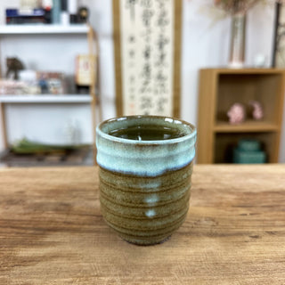 Die Teetasse "Shinrin" ist oben hellblau und unten dunkelgrün glasiert. Sie steht auf einem alten Holztisch. Im Hintergrund kann man verschwommen eine japanische Kalligraphie sowie zwei Regale mit Dekorationsobjekten erkennen.