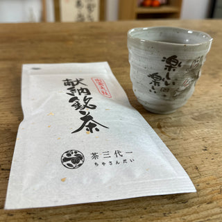 Die hellgraue Teetasse "Usagi", auf der man das herzige Motiv mit kleinen Hasen und japanischen Schriftzeichen sehen kann, steht auf einem Holztisch. Daneben liegt einen weiße Packung "Izumo Taisha Sencha", auf deren Vorderseite eine japanische Kalligraphie zu sehen ist.