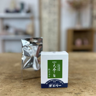 Im Vordergrund steht die weiße Schachtel "Tenko no Shiro" Matcha mit einem grünen Viereck auf der Vorderseite und einer dunkelblauen Basis. Dahinter steht ein silbriges Säckchen, der Inhalt der Verpackung.