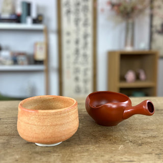 Auf einem Holztisch steht der Chawan "Satsuma" in Orange neben dem Abhkühlgefäß "Yuzamashi Tetsu" in rostrot. 