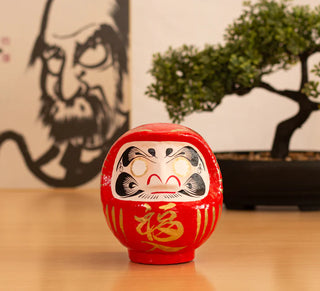 Japanische Daruma-Figur als Glücksbringer auf Holztisch, im Hintergrund Bonsai und Kalligrafie-Kunst