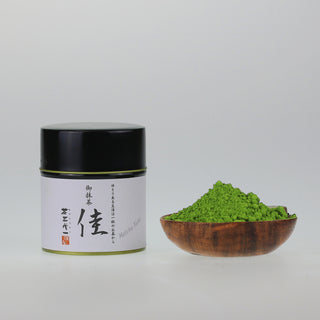 Matcha Yoshi in edler schwarzer Dose mit japanischer Kalligraphie, neben einer kleinen Holzschale mit grünem Matchapulver